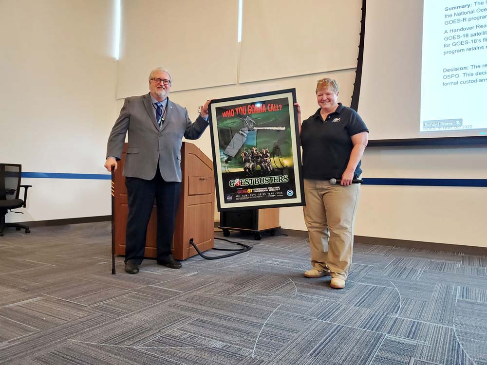 Two people standing in a conference room in front of a podium, holding a large, framed poster between them.